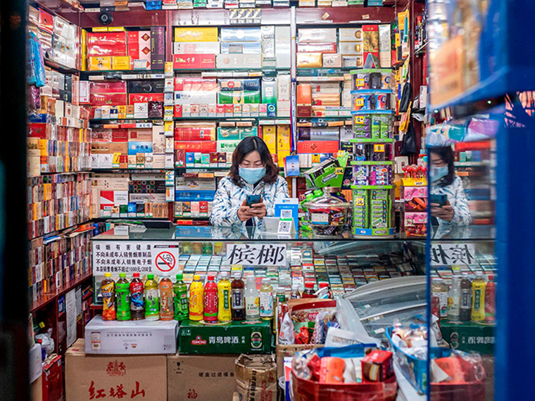 A masked person looks at their phone and sits behind a glass counter