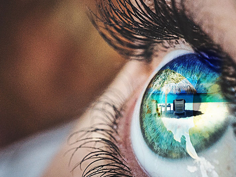 Close-up angled image of someone's eye. The eye is reflecting a beach landscape