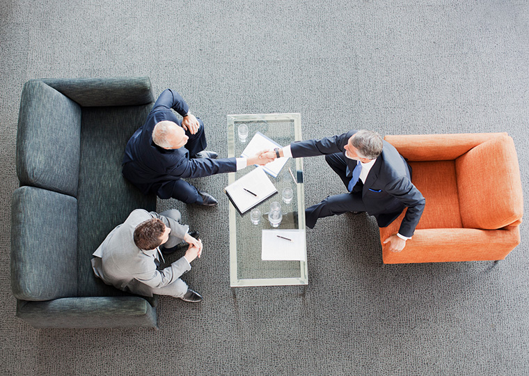 An overhead photo of two gentlemen on love seat one of whom is shaking hands with a man in an orange chair